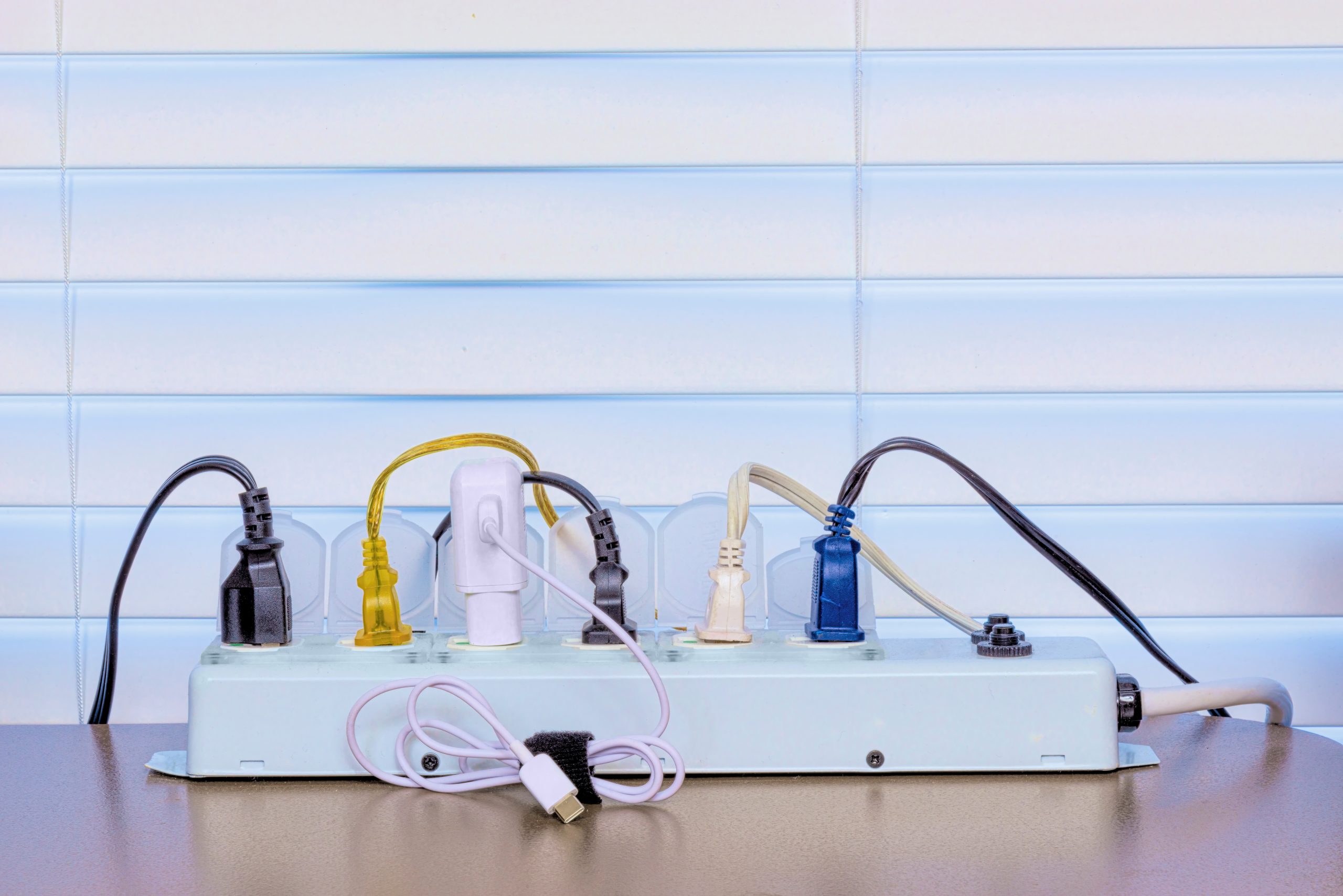 Horizontal shot of a full power strip on a desk in front of a Venetian blind background.